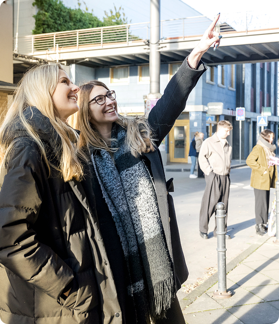 Zwei Frauen im Wintermantel stehen auf einem Bürgersteig, eine zeigt mit ausgestrecktem Arm nach oben