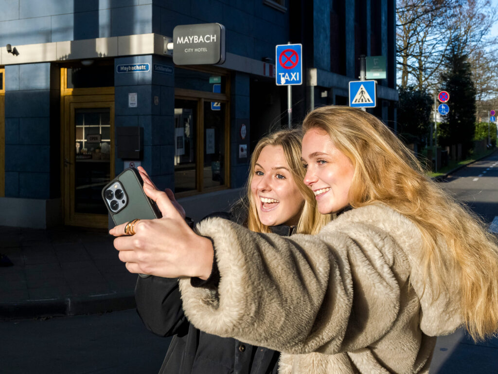 Zwei Personen mit langen Haaren machen ein Selfie mit einem Smartphone vor einem Gebäude