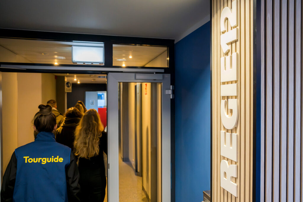 Person mit dunkelblauer Weste mit gelber Aufschrift 'Tourguide' von hinten vor mehreren Personen in einem Flur mit beleuchtetem Schild 'REGIE12' an der Wand