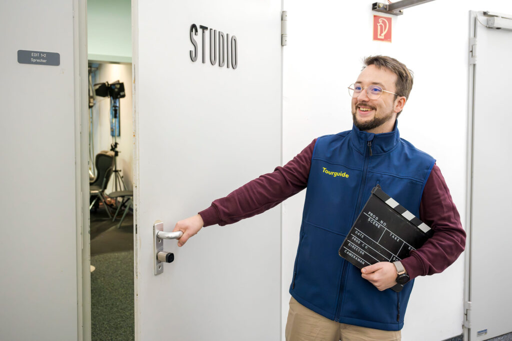 Person mit blauem Tourguide-Weste öffnet eine weiße Tür mit der Aufschrift 'STUDIO' und hält eine Filmklappe in der Hand